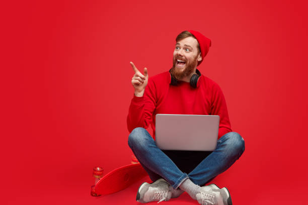Cheerful bearded hipster in vivid outfit pointing aside and smiling while using modern laptop and sitting near skateboard against red background
